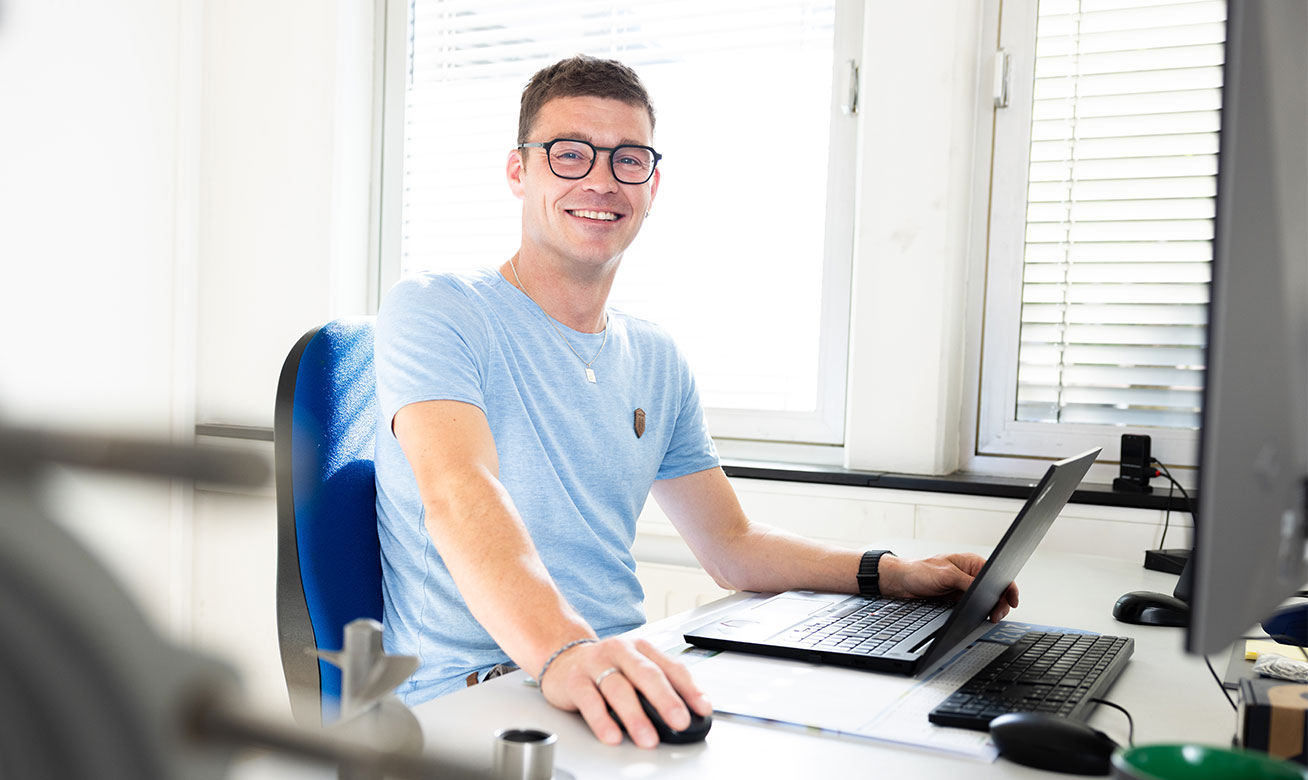 Ihr direkter Ansprechpartner im Customer Service Young man with glasses smiling at desk with laptop and computer in a bright office environment.