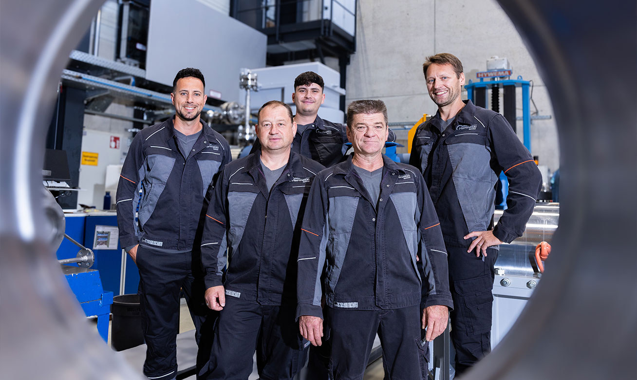 Field Service weltweit im Einsatz Five HERMETIC service technicians in uniform workwear posing together inside an industrial facility.