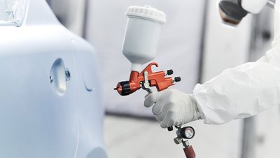 <h3>Production d'acide acrylique et d'acrylates</h3> Technician in white protective suit using an orange HVLP spray gun to paint a car body panel.