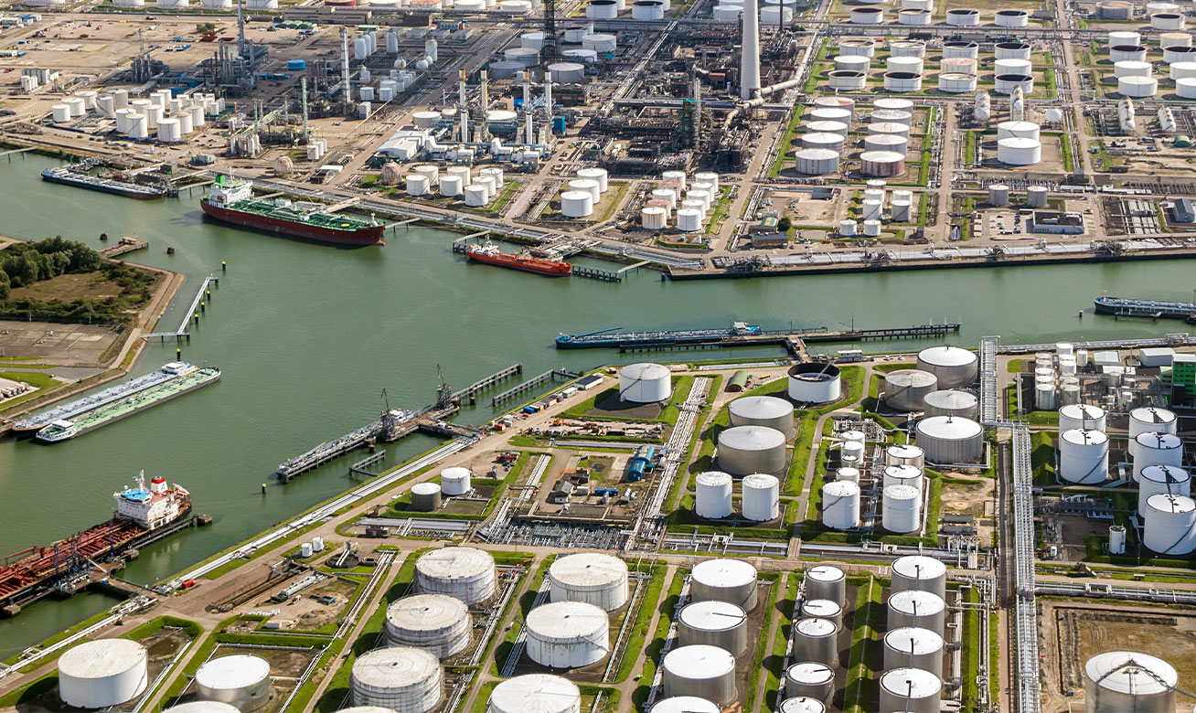 <h2>Transport en toute sécurité de fluides volatiles et inflammables</h2> Aerial view of a large petrochemical port with oil storage tanks, refineries, and tanker ships docked along industrial waterways.