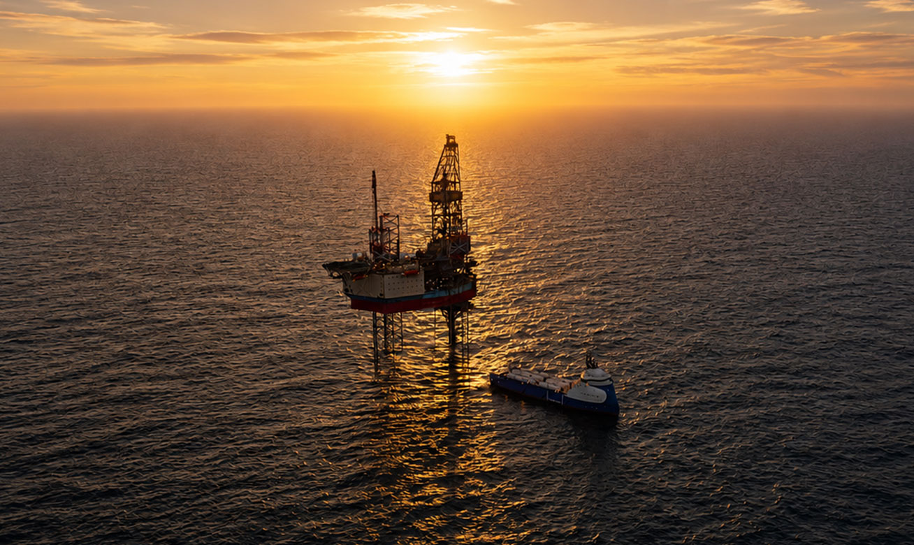 Offshore oil drilling platform and supply vessel at sunset over calm ocean waters, aerial view.