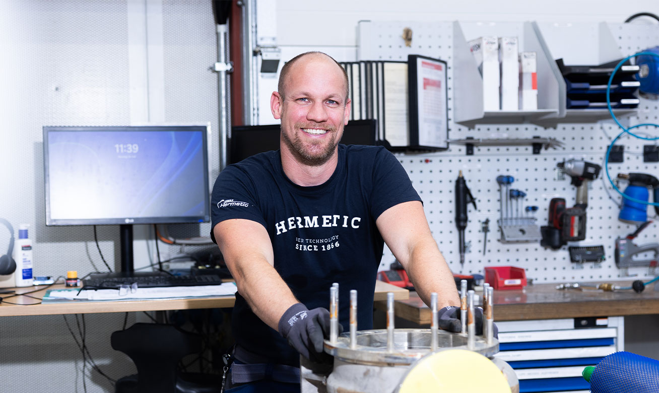 Reparaturservice für HERMETIC Pumpen Smiling technician in HERMETIC uniform working on mechanical equipment in a well-organized workshop with tools on pegboard.