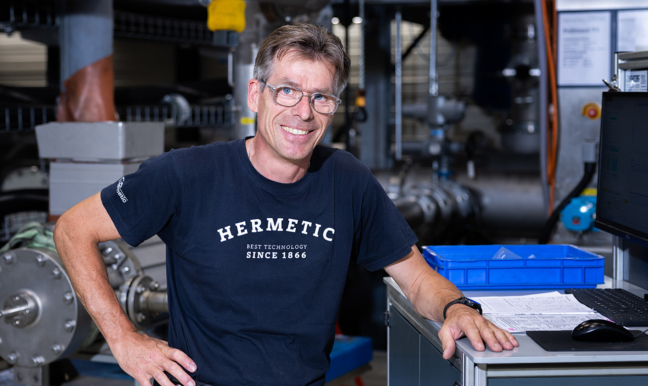 <h2>Fiabilité et pragmatisme </h2> Man in "HERMETIC Best Technology Since 1866" shirt smiling in an industrial facility with machinery and a workstation.