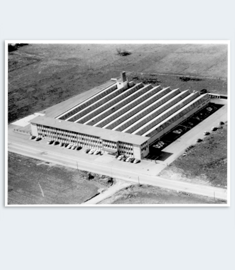 <h3>Déménagement à Gundelfingen</h3> Aerial black-and-white photo of a large industrial factory building with sawtooth roof, surrounded by fields and parking areas.