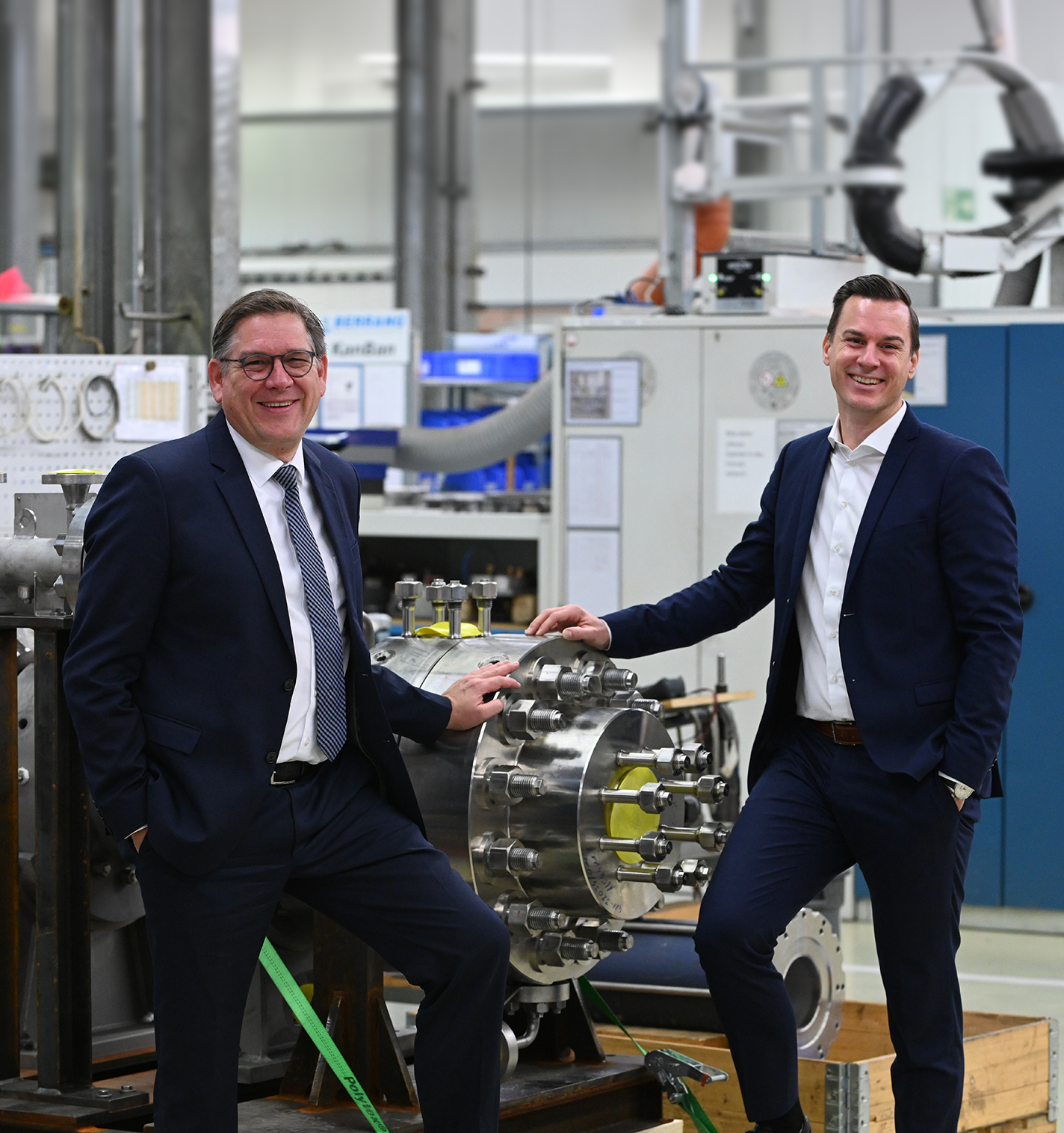 Two men in dark suits smiling in an industrial facility, posing with a metal valve or pump component.
