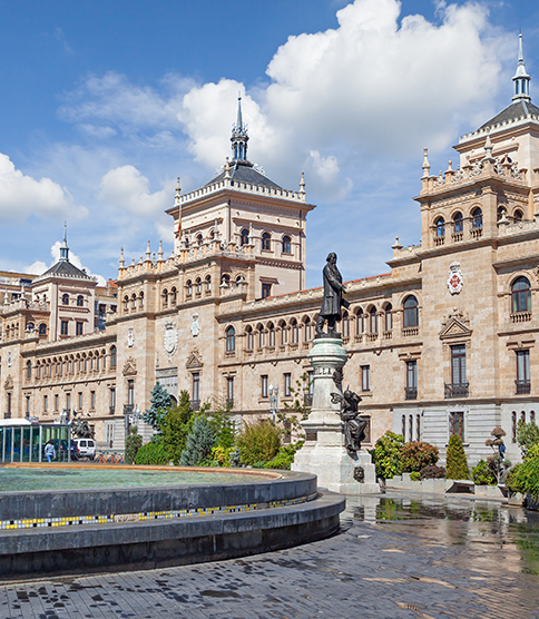 <h3>Ouverture de la succursale en Espagne</h3> Historic ornate building with towers and balconies, fountain and bronze statue in foreground plaza, blue sky above.