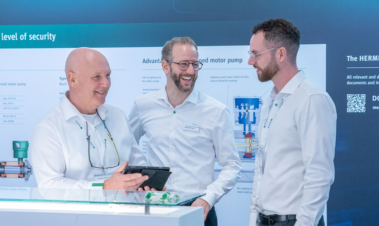 Three professionals in white shirts discuss at a trade show booth displaying canned motor pump technology.