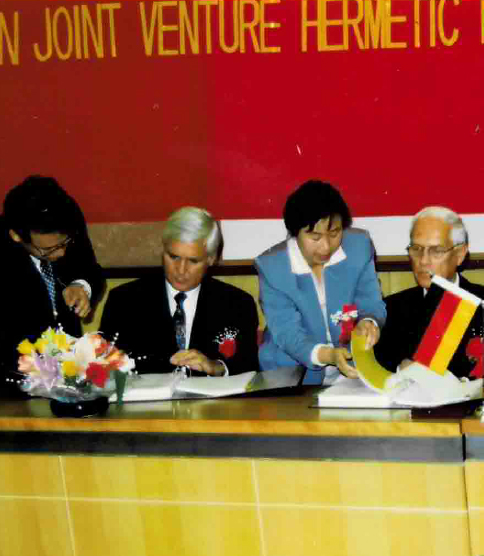 <h3>Coentreprise en Chine</h3> Signing ceremony at a Sino-German joint venture "HERMETIC," four people seated at a table with a German flag.