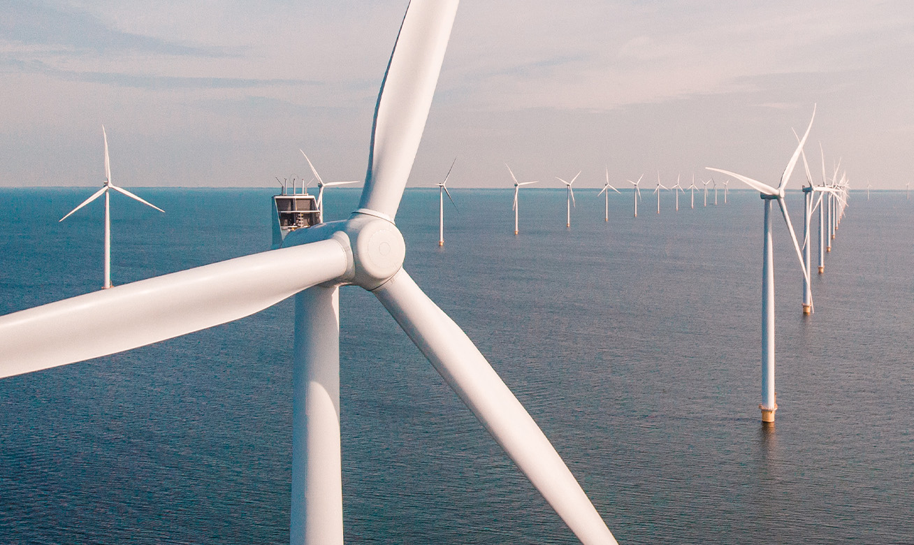Offshore wind farm with rows of white wind turbines standing in calm blue ocean water under a hazy sky.