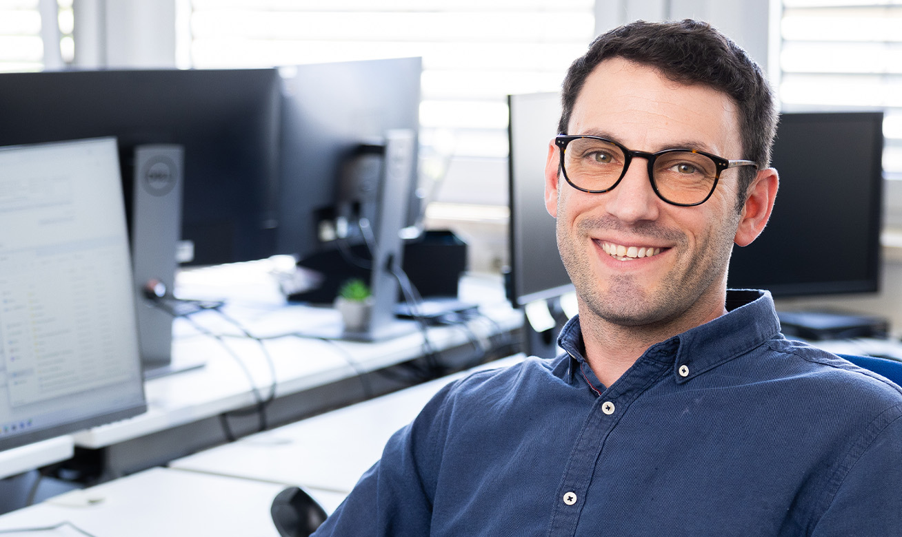 <h2>Système de gestion intégré</h2> Man with glasses smiling at desk with multiple computer monitors in an office environment.