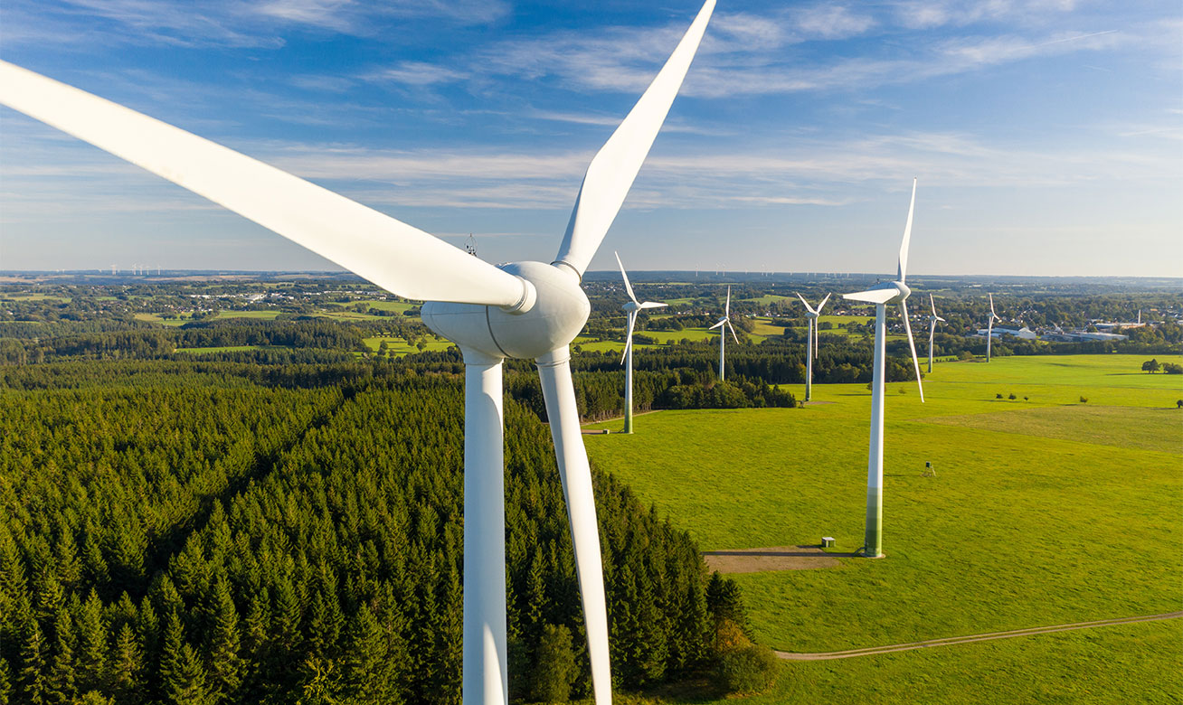 <h2>Un refroidissement efficace pour l'industrie, l'énergie et la mobilité</h2> Vue aérienne d'éoliennes dans un paysage verdoyant – gros plan sur un rotor au premier plan