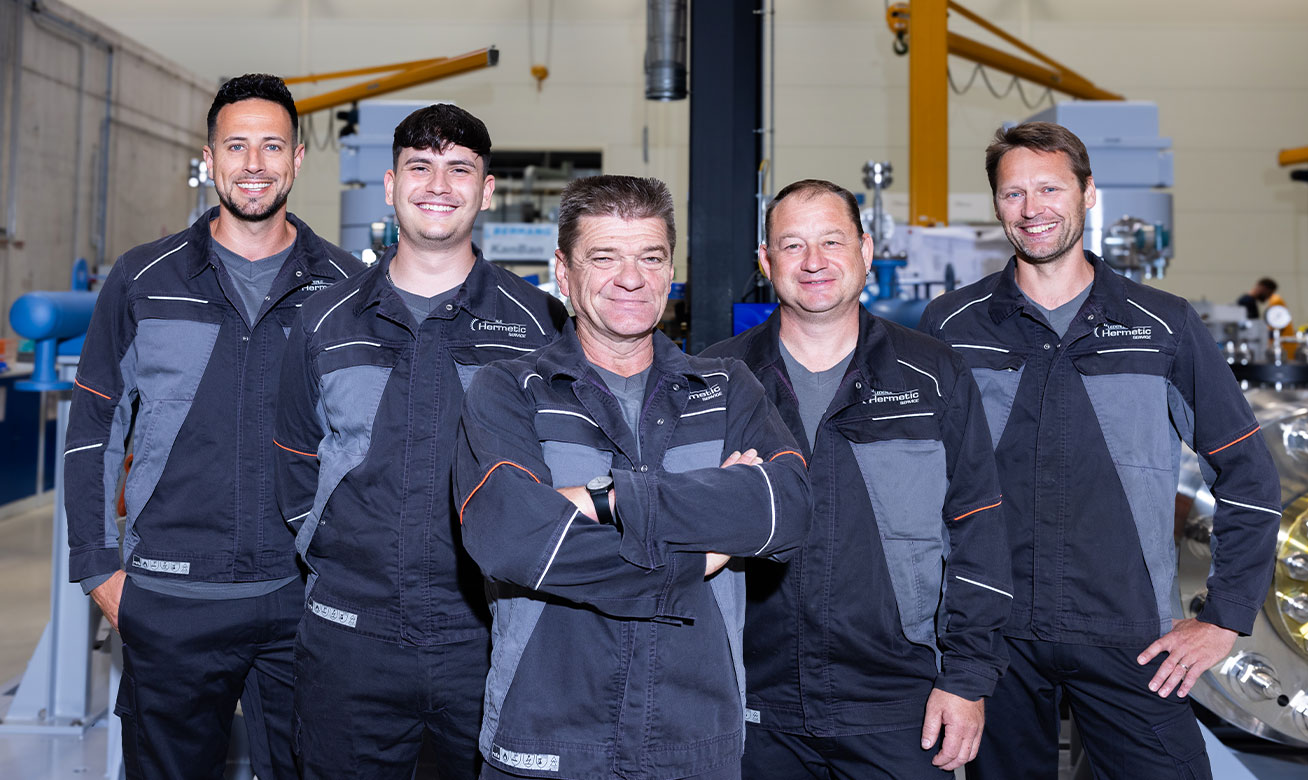 Five HERMETIC Service technicians in matching dark uniforms smile together inside an industrial manufacturing facility.