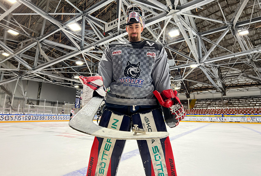 Hockey goalie in EHC Freiburg Wölfe jersey standing on ice rink, holding goalie stick, wearing full protective gear.