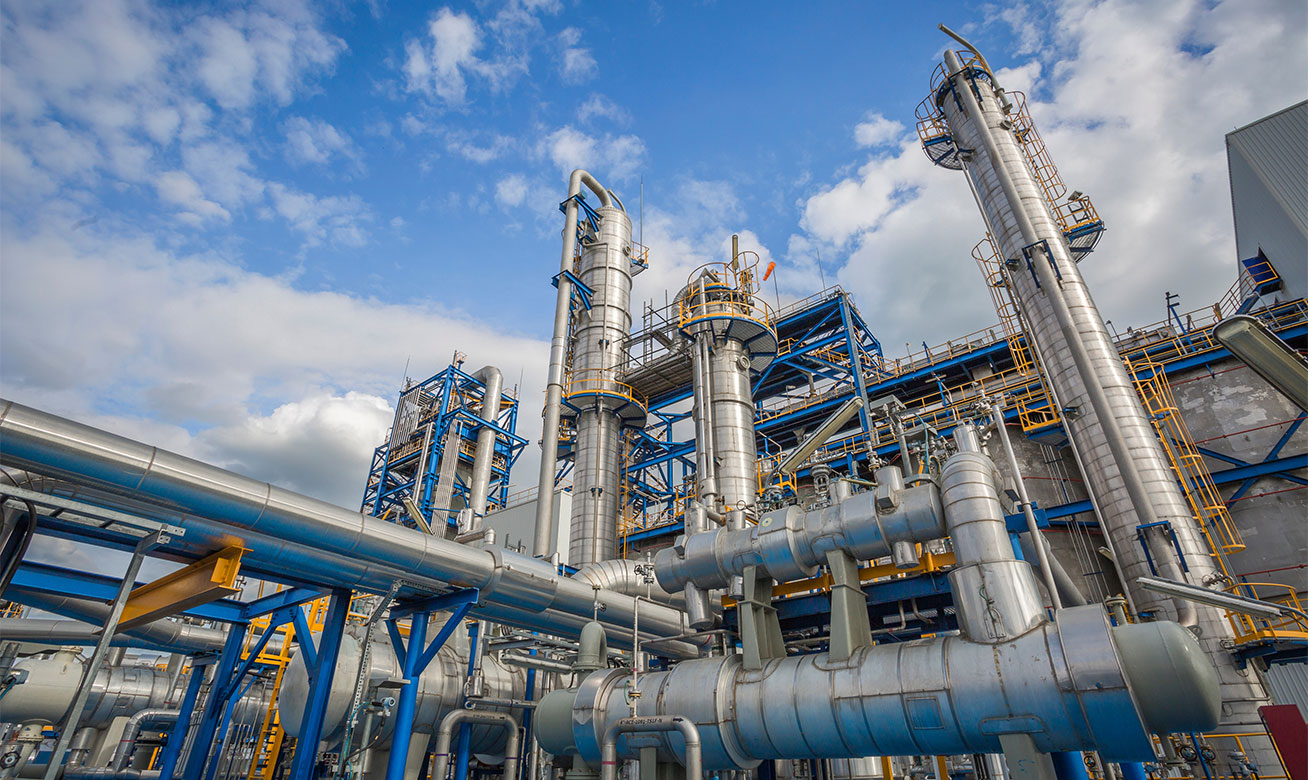 <h2>Solutions de pompes pour les processus exigeants du secteur pétrochimique</h2> Industrial oil and gas refinery with large silver pipes, distillation columns, and blue steel structures against a cloudy sky.