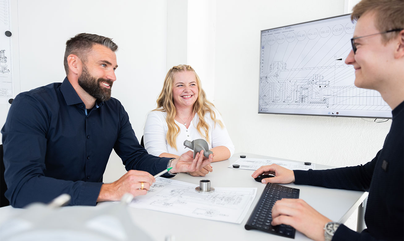 <h2>L'expertise au service de vos pompes</h2> Three engineers collaborate at a meeting table with technical drawings and a 3D-printed part, reviewing CAD schematics on a screen.