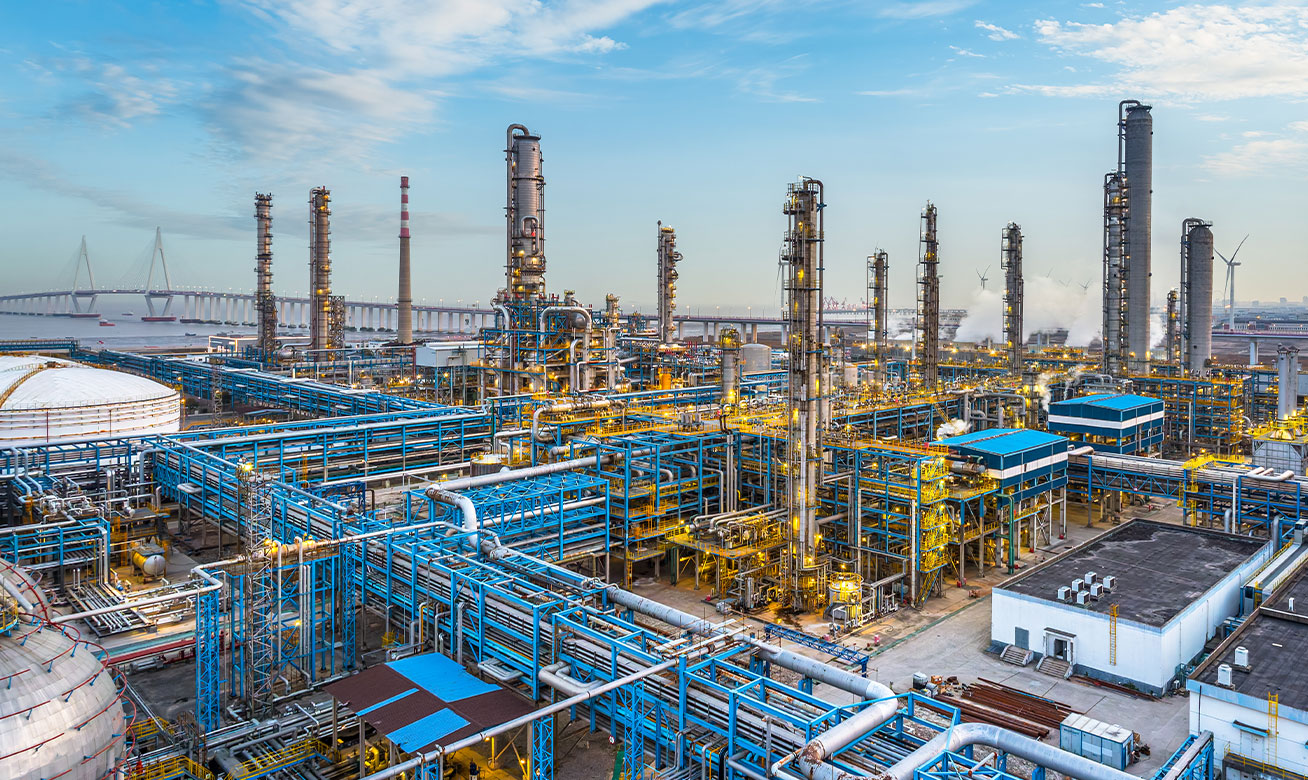 <h2>Manipulation en toute sécurité des intermédiaires réactifs</h2> Aerial view of a large petrochemical refinery with blue pipelines, distillation towers, and golden lights near a waterway.