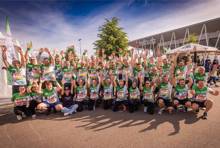 Group of runners in green team shirts with race bibs raising hands in front of SC Freiburg stadium